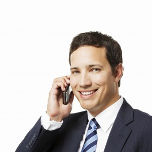Young businessman smiles at the camera while talking on his cellphone. Square shot.